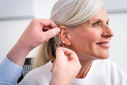 Ältere Frau bekommt ein modernes Hörgerät vom Hörakustiker angepasst und lächelt dabei.